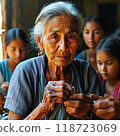 An elderly woman proudly teaches young girls traditional embroidery. Her weathered hands guide young girls, passing on cultural heritage in a simple village setting filled with warmth. 118723069