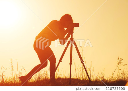 Silhouette of woman photographer shooting landscape with camera on tripod at sunset time in the field Silhouette of woman photographer shooting landscape with camera on tripod at sunset time in the field 118723600