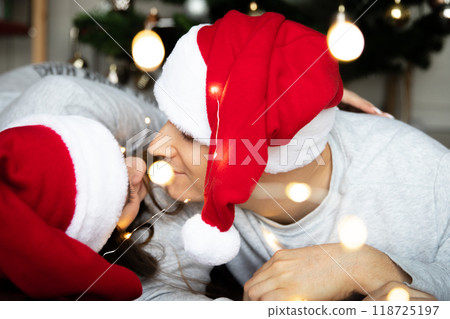 Young beautiful men women Santa hats looking each other tenderly near Christmas tree for New Year Young beautiful men women Santa hats looking each other tenderly near Christmas tree for New Year 118725197