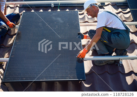 Workers building solar panel system on rooftop of house. Two men installers in helmets installing photovoltaic solar module outdoors. Alternative, green and renewable energy generation concept. 118728126