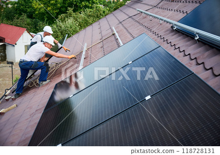 Workers building solar panel system on roof of house. Two men installers in helmets carrying photovoltaic solar module outdoors. Alternative, green and renewable energy generation concept. Workers building solar panel system on roof of house. Two men installers in helmets carrying photovoltaic solar module outdoors. Alternative, green and renewable energy generation concept. 118728131