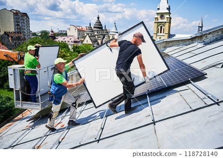 Workers building solar panel system on metal rooftop with assistance of crane lift in urban setting. Men installers carrying photovoltaic solar module outdoors. Renewable energy generation concept. 118728140