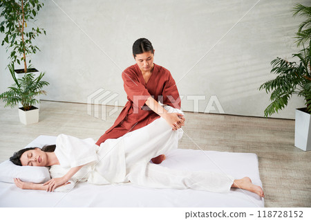 Masseuse making thai yoga massage. Woman therapist doing traditional massage treatment, stretching female patient's body. Client lying on mat, enjoying hand massage in calm atmosphere. 118728152