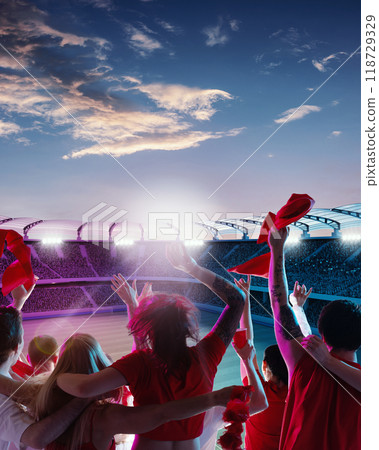 Group of fans in red shirts celebrating under colorful twilight sky, waving flags and scarves as field bathed in soft evening light. Group of fans in red shirts celebrating under colorful twilight sky, waving flags and scarves as field bathed in soft evening light. 118729329