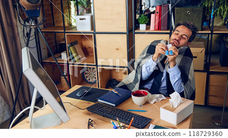 Employee tries to power through sickness at their desk, wrapped in a blanket, surrounded by tissues. Not taking sick days can lead to burnout. 118729360