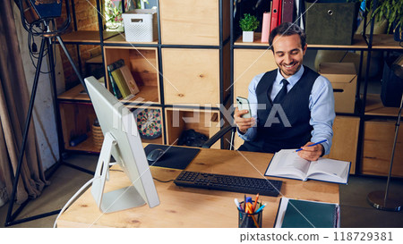 Businessman smiling while using his phone, scrolling through social media instead of focusing on work laid out in front of him. 118729381