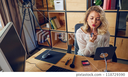 Female employee applies makeup at her desk. She's distracted from her work, emphasizing how personal tasks interfere with workflow and disrupt professional focus. 118729390