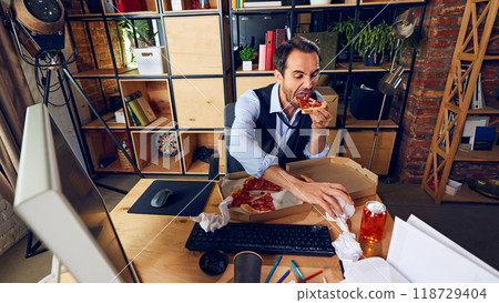 Businessman eating pizza while working. He wearing formal attire, but his eating habit reflects lack of professionalism in work setting. 118729404