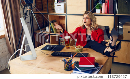 Beer in hand, feet on desk, young woman points at screen, seemingly frustrated while multitasking between work and casual drink. 118729437