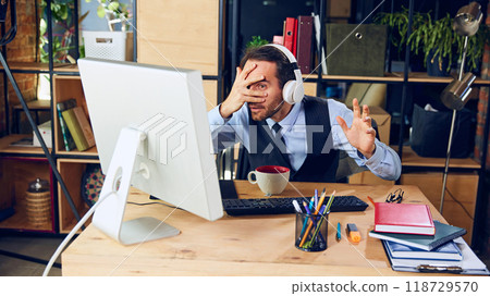 Overwhelmed by what's on screen, man covers his face, peeking through his fingers as he faces tricky work challenge. 118729570