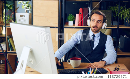 A young man dressed as office employee, wearing headphones at table with computer, looks at screen in surprise. Workspace. 118729571