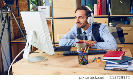 Young man dressed as office employee sitting in headphones at desk in office and looking at screen with interest. 118729573