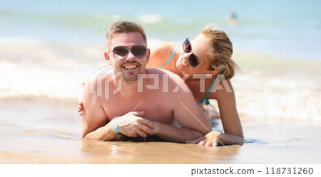 Cheerful couple resting together on beach laying on coastline and posing Cheerful couple resting together on beach laying on coastline and posing 118731260