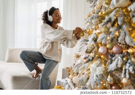 Merry Christmas. African American woman wearing headphones listening music decorating Christmas tree. Happy girl near classical traditional Christmas tree. Christmas eve at home time for celebration 118732572