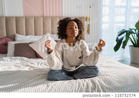 Yoga mindfulness meditation. Young healthy African girl practicing yoga at home. Woman sitting in lotus pose on bed meditating smiling relaxing indoor. Girl doing breathing practice. Yoga at home 118732574