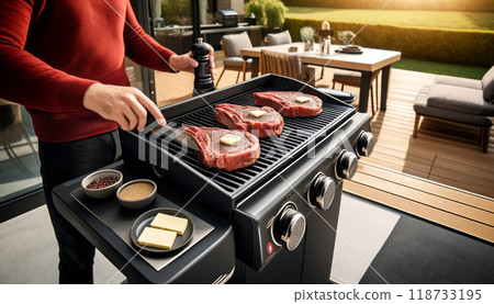 Essence of summer with a well-lit barbecue scene. A person in a light yellow shirt seasons steaks on a modern grill, surrounded by a lush garden under the bright afternoon sun. 118733195
