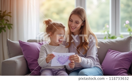 A mother and her young daughter are sitting on a sofa, reading a book together. The scene is cozy and warm, with a focus on family bonding. 118733632