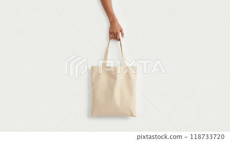 A person holds a plain beige shopping bag by the handle against a white background. The scene is minimalistic and focused on the bag. A person holds a plain beige shopping bag by the handle against a white background. The scene is minimalistic and focused on the bag. 118733720