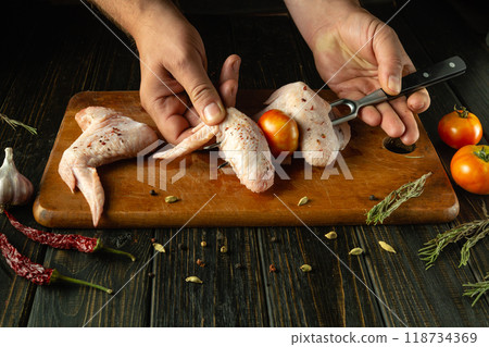 Chef prepares chicken wings with his hands for frying with spices on the grill. Preparation of cooking wings on the kitchen board. Fork in the hands of the cook Chef prepares chicken wings with his hands for frying with spices on the grill. Preparation of cooking wings on the kitchen board. Fork in the hands of the cook 118734369