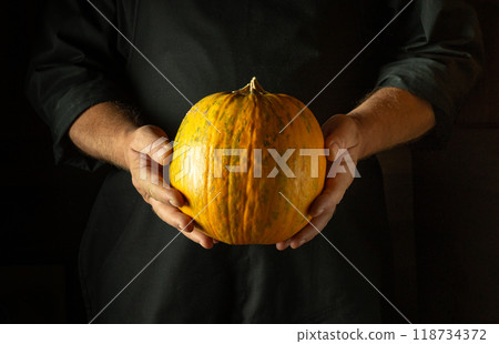 Chef holding a large ripe pumpkin before cooking or baking it in the kitchen. Low key concept of a fruit diet or cooking a national dish Chef holding a large ripe pumpkin before cooking or baking it in the kitchen. Low key concept of a fruit diet or cooking a national dish 118734372