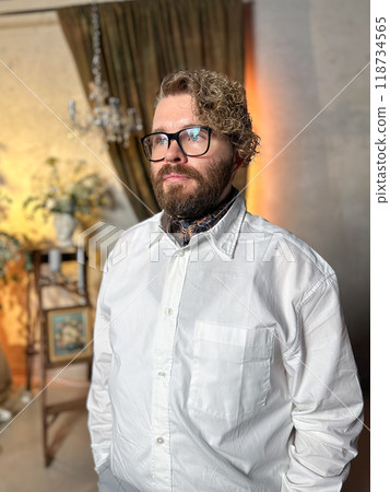 Thoughtful man in a white shirt stands confidently in a warmly lit room with a classic and elegant background interior. Confidence, elegance and male fashion concept Thoughtful man in a white shirt stands confidently in a warmly lit room with a classic and elegant background interior. Confidence, elegance and male fashion concept 118734565