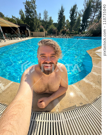 Millennial man taking selfie by swimming pool. Vibrant blue water and relaxed setting create a perfect atmosphere for summer fun. Holidays and vacation concept Millennial man taking selfie by swimming pool. Vibrant blue water and relaxed setting create a perfect atmosphere for summer fun. Holidays and vacation concept 118735840