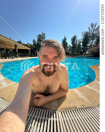 Millennial man taking selfie by swimming pool. Vibrant blue water and relaxed setting create a perfect atmosphere for summer fun. Holidays and vacation concept 118735850