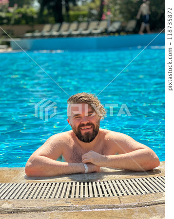 Millennial man enjoying a refreshing day by swimming pool, leaning on the pool's edge and beaming with a broad smile. Vibrant blue water and relaxed setting create a perfect atmosphere for summer fun 118735872