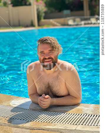 Millennial man enjoying a refreshing day by swimming pool, leaning on the pool's edge and beaming with a broad smile. Vibrant blue water and relaxed setting create a perfect atmosphere for summer fun Millennial man enjoying a refreshing day by swimming pool, leaning on the pool's edge and beaming with a broad smile. Vibrant blue water and relaxed setting create a perfect atmosphere for summer fun 118735889