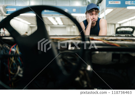 Tired mechanic fixing car in repair garage, frustrated but focused, surrounded by tools and equipment. Pensive repairman in car service looking at car thinking of how to repair in garage car service. 118736463