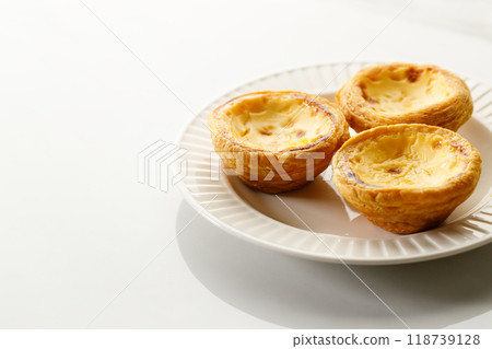 Stack of Portuguese sweet dessert egg tart or Pastel de Nata on white ceramic plate on white table. Copy space. Selective focus. Stack of Portuguese sweet dessert egg tart or Pastel de Nata on white ceramic plate on white table. Copy space. Selective focus. 118739128