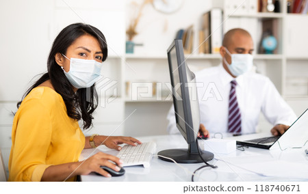 Confident Latina in medical mask working on computer in office 118740671