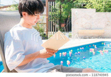 年輕的亞洲男子坐在泳池邊的躺椅上看書 年輕的亞洲男子坐在泳池邊的躺椅上看書 118740945
