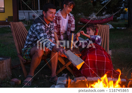 Family is sitting by the outdoor campfire in the courtyard of the house on camping chairs, drink tea from a thermos, warm up and chat by the hearth Family is sitting by the outdoor campfire in the courtyard of the house on camping chairs, drink tea from a thermos, warm up and chat by the hearth 118745865