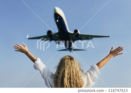 A woman with her arms raised above her head waves at an airplane flying over her 118747815