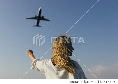 A woman with her arms raised above her head waves at an airplane flying over her 118747816
