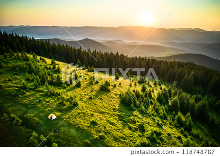 Aerial view of campsite on grassy hillside, with tents scattered amidst lush greenery and small trees. Golden hues of sunrise. In distance, layers of rolling mountains fade into horizon. 118748787