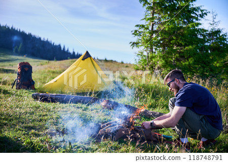 Man tending to campfire on grassy field with yellow tent and backpack nearby. Smoke rises from fire as he works, surrounded by lush greenery and distant tree line under clear blue sky. 118748791