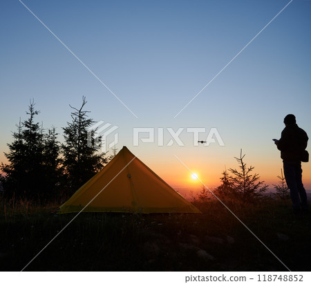 Peaceful campsite at sunrise with vibrant yellow tent pitched among pine trees. Silhouette of person operating drone adds dynamic element to serene scene, as sun casts golden glow across horizon. 118748852