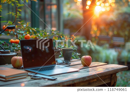 A calm garden workspace features a laptop, apples, and a steaming cup of coffee as the sun sets, casting a warm glow 118749061