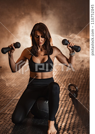 An empowered woman confidently lifts weights in a vibrant fitness studio, showcasing her strength, commitment, and dedication to fitness and wellbeing through various exercise forms 118752845