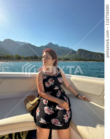 Woman in a black dress sits on a boat under the bright sun, with a scenic view of mountains and the sea in the background. Moment of tranquility and natural beauty concept. Vacation, travel and voyage 118756880