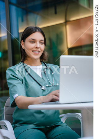 Smiling pretty young female doctor sitting at the table and looking confident Smiling pretty young female doctor sitting at the table and looking confident 118758228