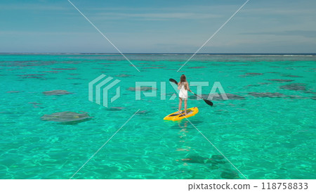 Woman paddleboarding in tropical waters, enjoying vacation in paradise. Crystal-clear turquoise sea, sunny sky, and coral reefs visible below. Perfect getaway for relaxation and fun 118758833