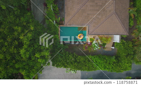 Aerial view of a woman enjoying a sunny day, floating on a yellow inflatable ring in a private pool surrounded by lush tropical vegetation Aerial view of a woman enjoying a sunny day, floating on a yellow inflatable ring in a private pool surrounded by lush tropical vegetation 118758871