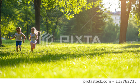 Two energetic kids, boy and girl, sprint across a lush green park bathed in warm sunlight, embodying the freedom and joy of outdoor play 118762733