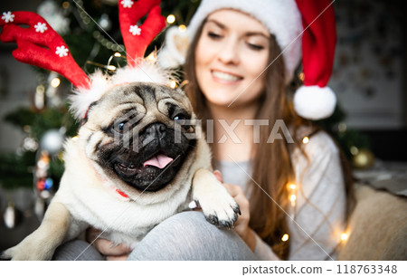 Funny cute pug dog in red Christmas reindeer horns sitting in arms of a beautiful girl in Santa hat. 118763348