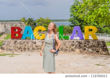 Female tourist posing in front of the colorful BACALAR sign with vibrant letters, trees, and Bacalar Lake in the background. Mexican travel and adventure concept 118766021