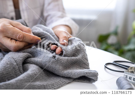 A woman uses her hands to sew up a hole in a gray knitted garment with a needle and thread.  118766173