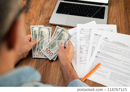 Man counting cash while reviewing tax documents on a wooden desk. Financial management, tax refund, and accounting concept.  118767125